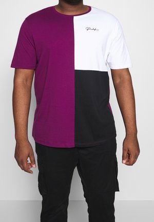 Mann trägt ein kurzärmliges T-Shirt mit Farbfeldern in Lila, Weiß und Schwarz, kombiniert mit schwarzer Hose vor einem einfarbigen Hintergrund.