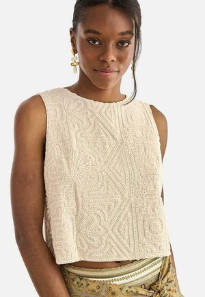 Mujer con top beige sin mangas con bordado geométrico en relieve y falda estampada, posando sobre fondo blanco liso.