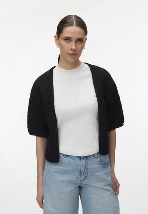 Kobieta z gładko zaczesanymi włosami, nosząca srebrne kolczyki koła, teksturowany czarny krótki sweter na białej koszuli i jasnoniebieskie dżinsy.