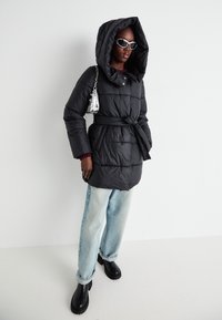 Zwarte pufferjack met een grote capuchon, een tailleband en een gewatteerd patroon. Gecombineerd met lichtblauwe jeans en zwarte chunky laarzen. Inclusief zilveren handtas.