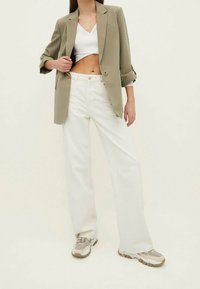 Blazer verde oliva, top corto bianco, pantaloni larghi color crema e scarpe sportive beige; linee pulite, vestibilità rilassata e design minimalista.