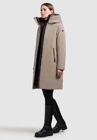 Beige vandtæt parka med høj krave, lynlås foran og to sidelommer. Sammen med sorte leggings og knæhøje sorte støvler.