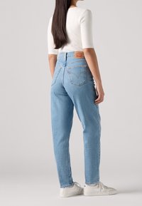 Jean taille haute bleu clair avec une coupe décontractée, présentant une patch en cuir de marque, des poches arrière et des ourlets retroussés, assortis à des baskets blanches.