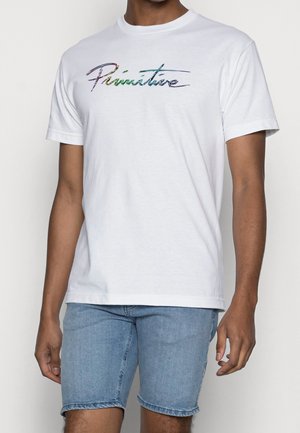 Uomo che indossa una maglietta bianca con la scritta colorata "Primitive" e pantaloncini di jeans azzurri su uno sfondo semplice.