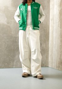 Chaqueta varsity verde y blanca con botones, que presenta un logo de "C"; combinada con pantalones blancos holgados de textura y zapatos beige.
