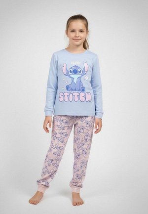 Jeune fille portant un haut bleu à manches longues avec le personnage "Stitch" et un pantalon rose orné de plusieurs visages de Stitch, debout pieds nus.