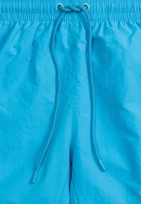 Jasnoniebieskie kąpielówki wykonane z lekkiego materiału, z elastyczną talią i sznurkiem do regulacji. Gładka faktura bez widocznych wzorów.