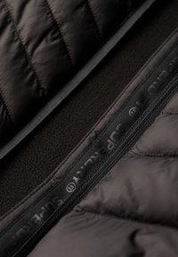 Capa exterior acolchada negra, interior de suave forro polar negro, cremallera visible con marca, costuras planas y sutiles variaciones de textura.