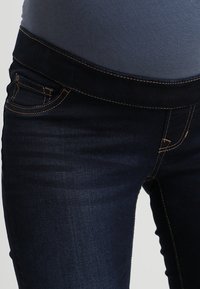 Jeans de mezclilla de un azul oscuro con una textura suave, que presentan una cinturilla alta elástica, bolsillos frontales y costuras doradas en contraste.
