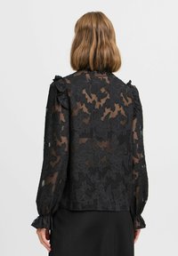 Schwarze durchsichtige Bluse mit floraler Stickerei, langen Ärmeln, Rüschendetails an den Schultern und gerafften Manschetten. Glatte Textur und leichtes Material.