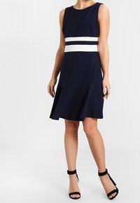 Námořnické bezrukávové šaty s bílým horizontálním pruhem v pase. A-line střih se hladkým materiálem a jemným rozšířením na lemu.