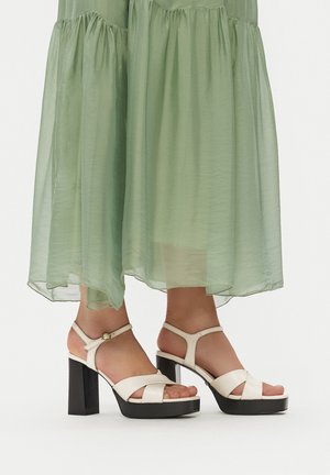 Sandalias de tacón alto blancas con tiras y plataforma negra gruesa, colocadas sobre un vestido verde transparente, mostrando las correas y los accesorios en el tobillo.