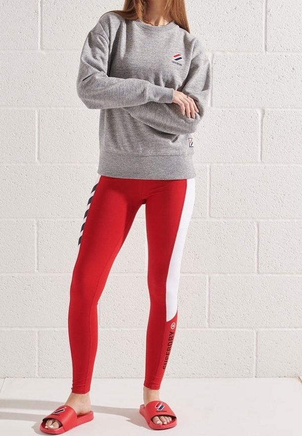 Femme portant un sweat-shirt gris, des leggings rouges et blancs avec le texte « Superdry » et des sandales rouges, debout contre un mur en briques blanches.