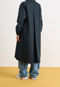 Czarny długi płaszcz z plisowanym wzorem, wykonany z gładkiego materiału. Zestawiony z oversize'owymi jasnoniebieskimi dżinsami i białymi trampkami z różowymi akcentami.