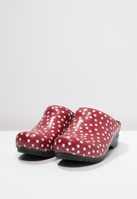 Zoccoli rossi realizzati in materiale liscio, con pois bianchi, punta arrotondata e suola nera. Esposti con un leggero angolo.