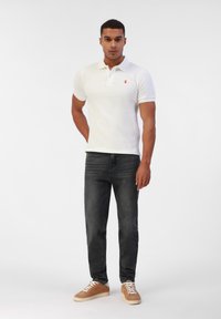 Polo blanc à manches courtes avec un petit emblème rouge, associé à un jean gris foncé et des baskets beiges. Design simple et épuré.