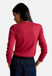 Kobieta z długimi ciemnymi włosami, ubrana w czerwoną bluzkę z długimi rękawami i ciemnoniebieskie dżinsy, widziana z tyłu na jednolitym tle.