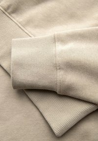 Beige katoenen trui stof met geribbelde manchetten, een gladde textuur en stikseldetails. De manchetten zijn lichtjes omgevouwen over het hoofdgedeelte.