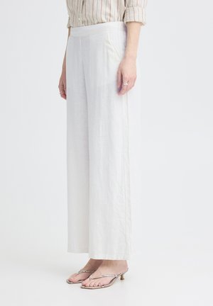 Kvinde iført hvide bukser med vide ben, stribet skjorte med opkaldte ærmer og kitten heel sandaler på en ensfarvet hvid baggrund.