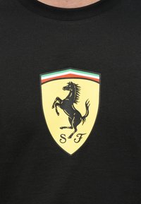 Κίτρινο λογότυπο Ferrari σε σχήμα ασπίδας με μαύρο άλογο που πηδά και πράσινες, λευκές και κόκκινες ρίγες σε μαύρο υφασμάτινο φόντο.