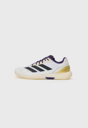 White athletic shoe with black stripes, purple accents, and gold heel detail. Features a textured sole and breathable mesh upper.