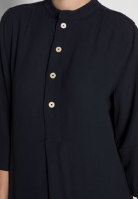 Blusa blu navy con colletto alla mandarina, maniche a tre quarti e cinque bottoni beige sul davanti. Tessuto dalla texture liscia.