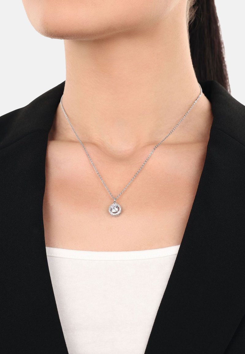 Collana d'argento con un pendente circolare che presenta una pietra centrale circondata da pietre più piccole, indossata sopra una maglietta bianca e una giacca nera.