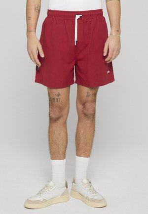 Mann som har på seg røde shorts med snor, hvite sokker og hvite joggesko, stående mot en enkel lys grå bakgrunn.