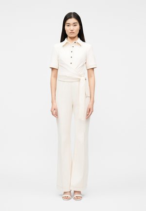 Mujer de pie vistiendo un mono corto de manga corta color crema con botones, cinturón envolvente, pantalones de pierna ancha y sandalias abiertas sobre un fondo blanco.