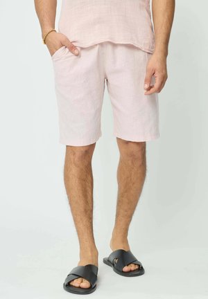 Des shorts en lin rose clair avec des poches latérales et une coupe décontractée. Portés avec des sandales noires, présentant un design de bride texturée.