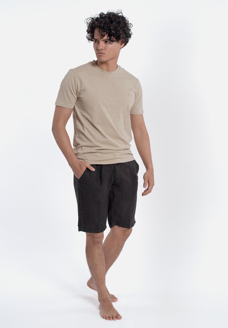 Beżowa koszulka z krótkim rękawem i okrągłym dekoltem, w połączeniu z czarnymi szortami. Model stoi boso, z rękami w kieszeniach szortów, prezentując swobodny wygląd.