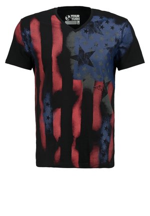 Homme portant un t-shirt noir, rouge et bleu à motifs d'étoiles, un short cargo blanc et des baskets noires, marchant sur un fond clair uni.