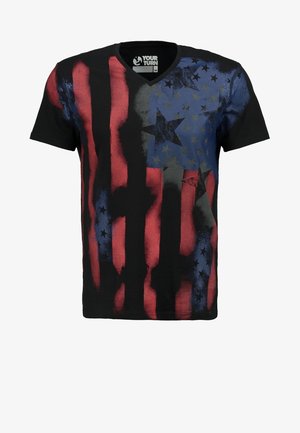 Homme portant un t-shirt noir, rouge et bleu à motifs d'étoiles, un short cargo blanc et des baskets noires, marchant sur un fond clair uni.