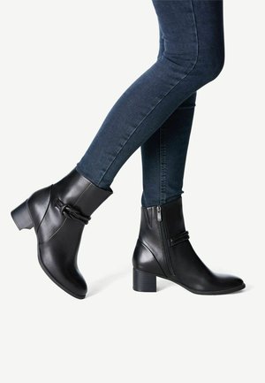 Person, die eng anliegende dunkelblaue Jeans und schwarze Stiefeletten mit niedrigen Blockabsätzen und seitlichen Reißverschlüssen vor weißem Hintergrund trägt.