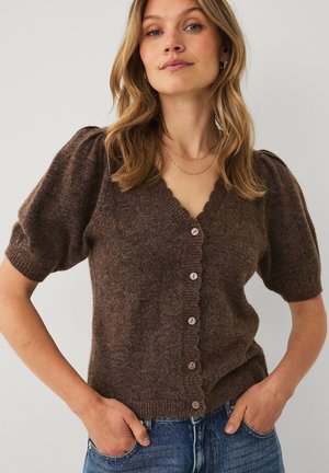 Kvinde iført en brun knappet kortærmet sweater med pufskuldre og blå jeans, stående med hænderne i lommerne mod en ensfarvet baggrund.
