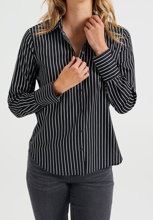 Vrouw draagt een zwart blouse met witte verticale strepen en donkergrijze jeans, terwijl ze met beide handen haar kraag rechtzet.