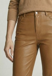 Persoon draagt nauwsluitende lichtbruine leren broek en een lichtbruine gebreide trui, hand naast dij rustend, neutrale achtergrond.