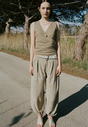 Jeune femme portant un haut beige sans manches et un pantalon beige ample, debout sur un chemin ensoleillé entouré d'arbres et d'herbe sèche.