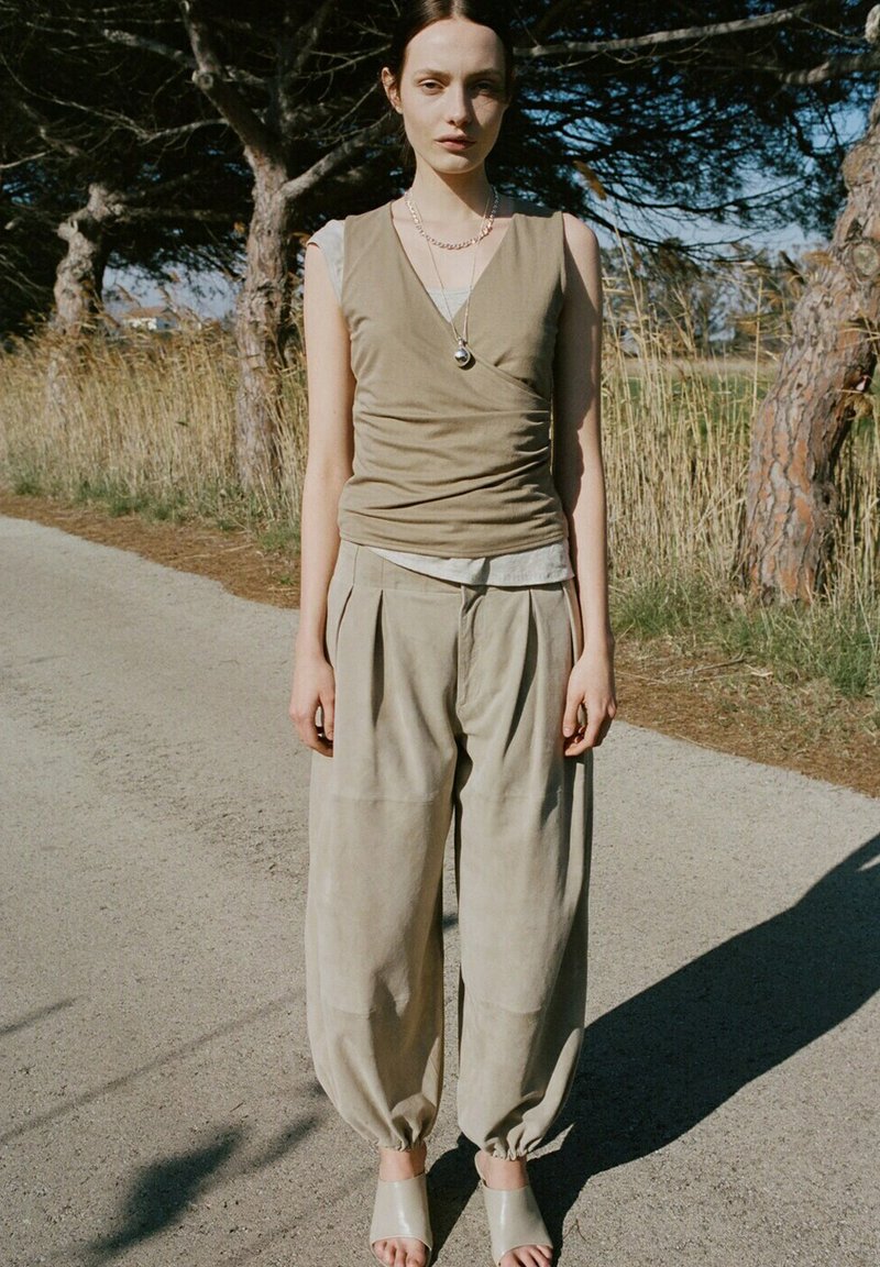 Jeune femme portant un haut beige sans manches et un pantalon beige ample, debout sur un chemin ensoleillé entouré d'arbres et d'herbe sèche.