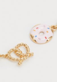 Auksinės spalvos metalinė pakabuko kaklo grandinėlė, turinti susuktos pretzel formos ir rožinį donutą su spalvingais barstytuvais. Švelnūs ir blizgūs paviršiai.