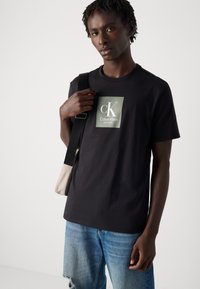 Joven vestido con una camiseta negra de Calvin Klein y jeans azules rasgados, sosteniendo un bolso beige de hombro con una correa negra.