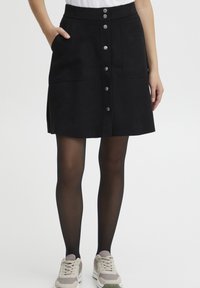 Jupe en daim noir avec une taille cintrée, fermeture à boutons sur le devant et poches latérales. Portée avec des collants noirs et des baskets grises.