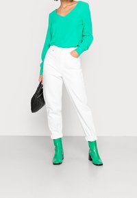 Grünes Langarmshirt mit V-Ausschnitt, weiße hochgeschnittene Hose und grüne Knöchelstiefel. Eine schwarze strukturierte Handtasche ergänzt das Outfit.