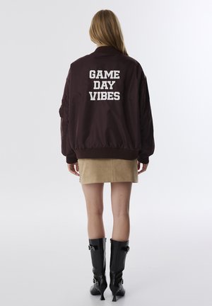 Bruine bomberjack met ribgebreide manchetten en zoom, voorzien van witte geborduurde tekst "GAME DAY VIBES" op de achterkant. Gepaard met een tan rok en zwarte laarzen.
