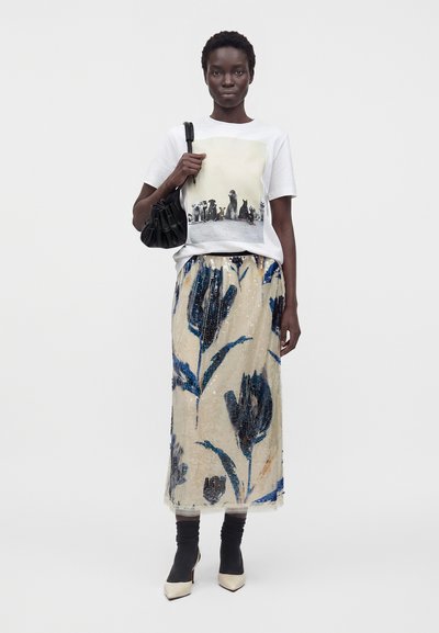 Femme portant un t-shirt blanc avec un imprimé chien, une longue jupe beige avec un motif floral bleu, des chaussettes noires, des talons blancs, et tenant un sac à épaule noir.