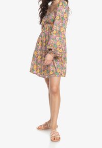 Robe à imprimé floral avec manches longues bouffantes, corsage froncé et jupe évasée dans des couleurs vives. Portée avec des sandales à enfiler rose clair.