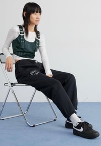 Grönrandig kort topp med justerbara band, svarta sweatpants med logotyp och svarta sneakers, sittande på en silverfärgad hopfällbar stol mot en ljus bakgrund.