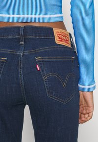 Tummansiniset farkut, joissa on nahkapatche, jossa lukee "Levi Strauss & Co." ja punainen lappu takataskussa. Keltaiset tikkaukset erottavat taskut.