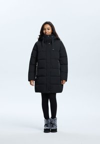 Zwarte pufferjas met hoge kraag, voorzien van een ritszak, trekkoordkap en quilted ontwerp. G gedragen met zwarte leggingen en fuzzy laarzen.