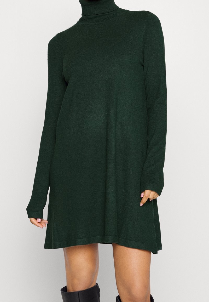 Robe tricotée à col roulé vert foncé à manches longues portée au-dessus des genoux avec des bottes noires mi-mollet.